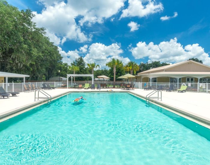 A person swims alone in a bright outdoor pool at a Bushnell RV resort in Central Florida, surrounded by lounge chairs, umbrellas, and trees, with a building and a blue sky filled with fluffy clouds in the background.