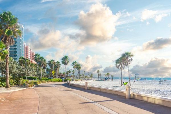 A sunny beachside promenade lined with palm trees and greenery, with high-rise buildings on the left and soft clouds in a blue sky over the sandy shore—perfect for those dreaming of a Central Florida escape or planning an RV resort stay.