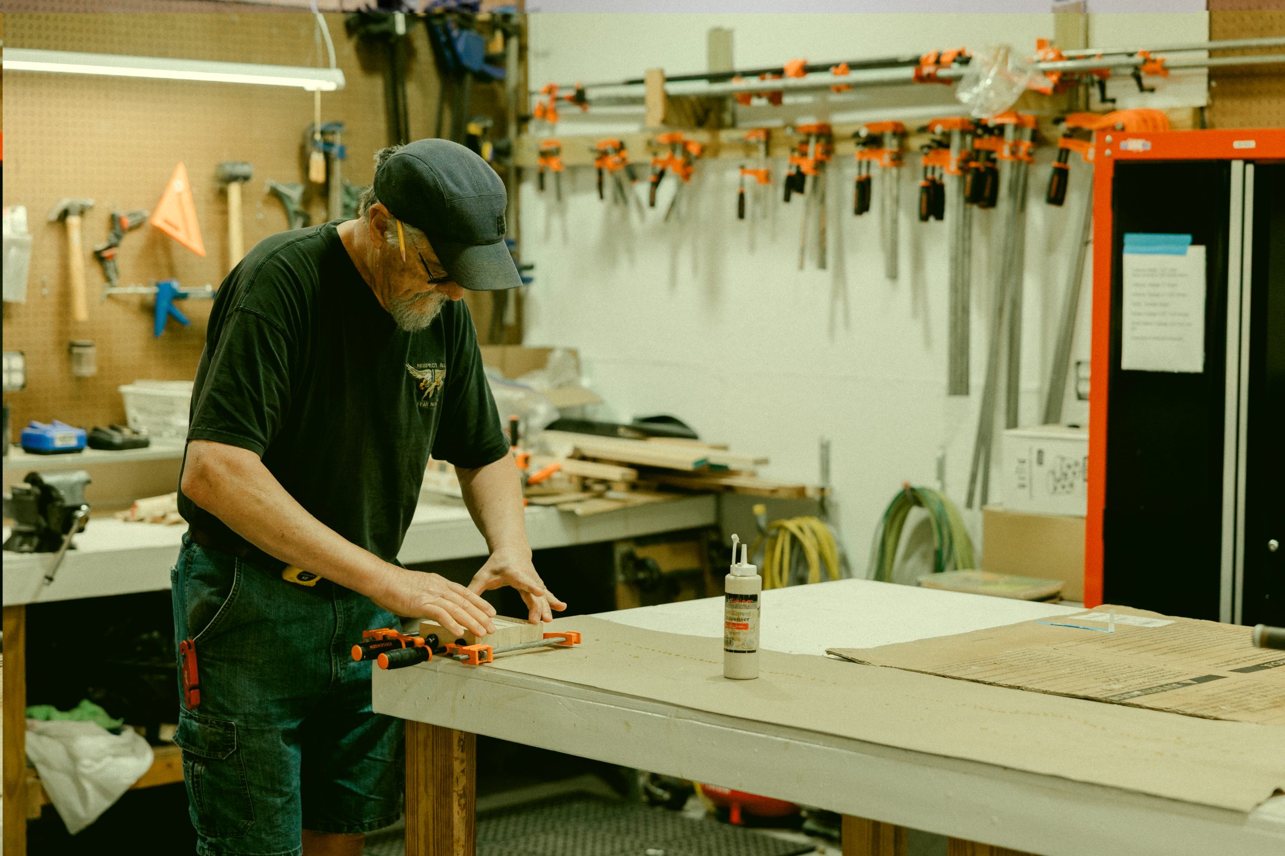 In a workshop near Central Florida, a person wearing a cap and glasses uses wood clamps at a table, surrounded by an array of tools—crafting with skill just steps from their RV resort in Bushnell.