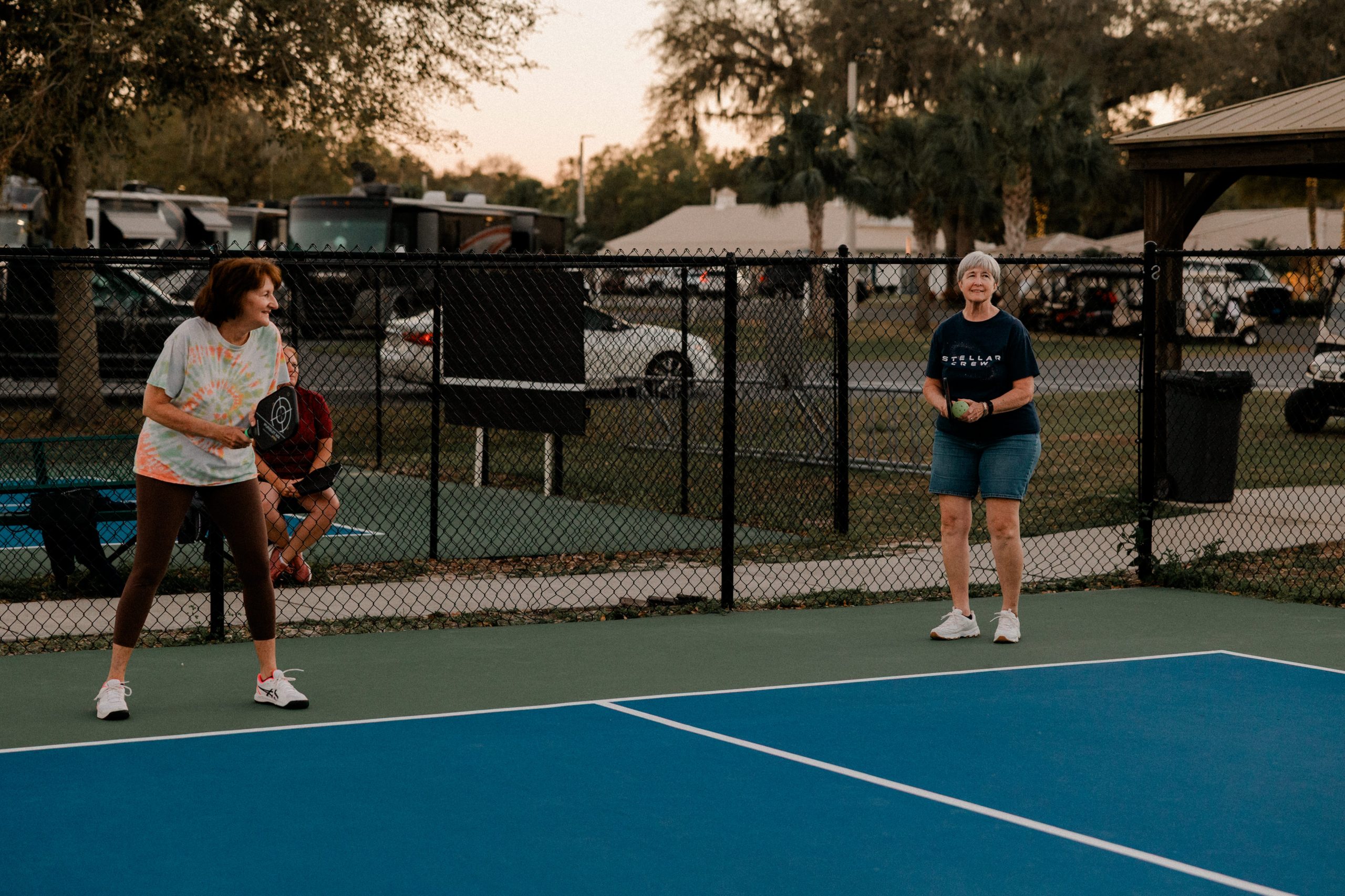 Two women stand on a pickleball court at Paradise Oaks RV in Bushnell, Central Florida, holding paddles and smiling, ready to play. It’s early evening with trees, parked cars, and the RV park visible in the background.