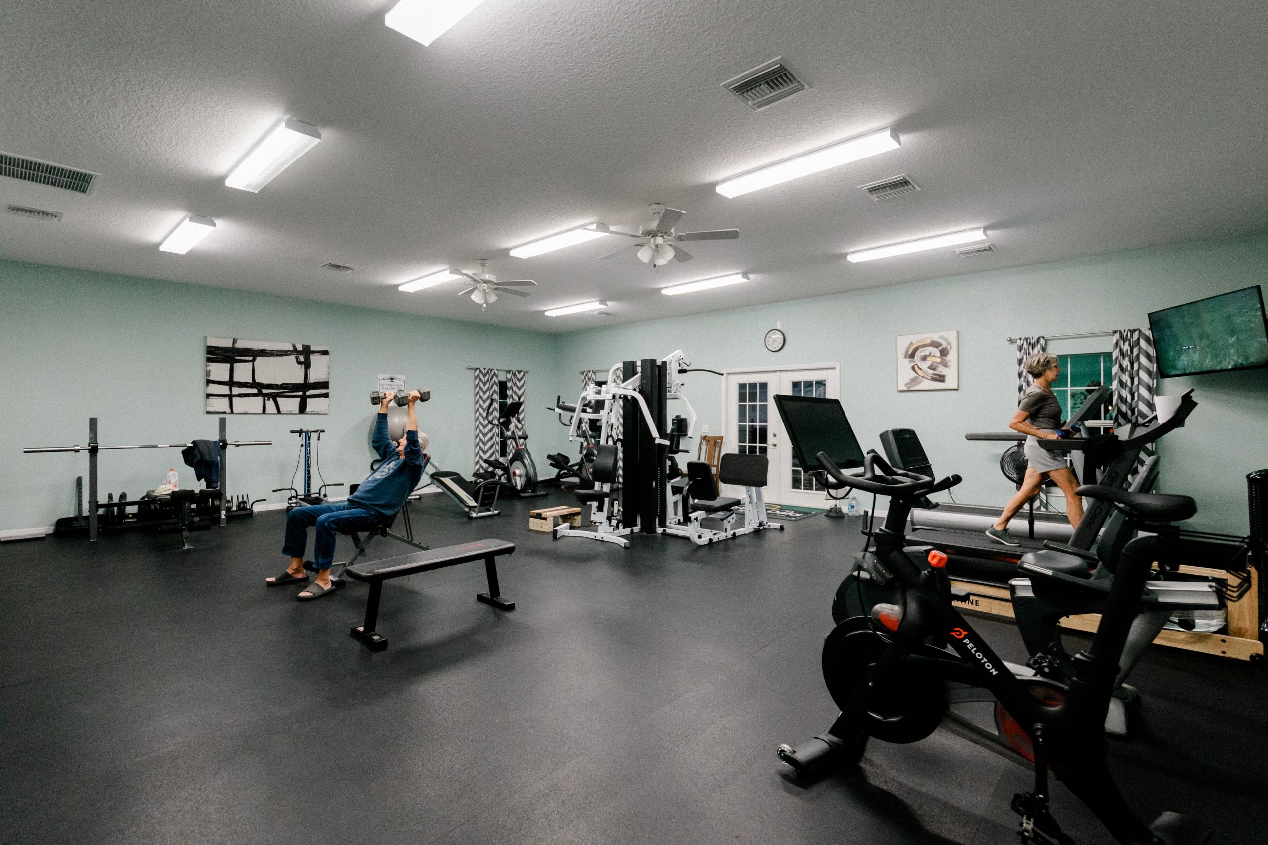 Two people exercise in a bright gym at Paradise Oaks RV resort; one lifts dumbbells on a bench while the other walks on a treadmill. The Central Florida facility features modern equipment, mint green walls, and vibrant ceiling lights.