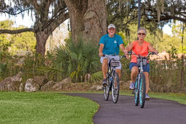An older man and woman ride bicycles on a paved path through a park at Paradise Oaks RV Resort in Central Florida, smiling and enjoying a sunny day surrounded by large trees and lush greenery.