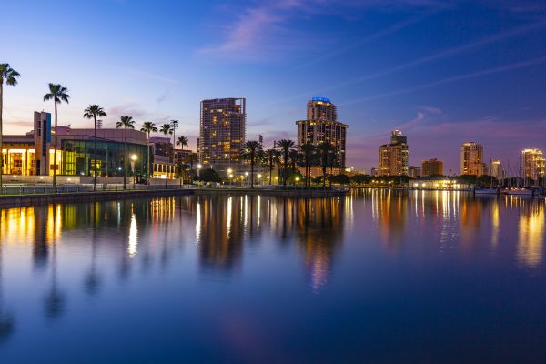 City skyline at dusk with tall buildings, palm trees, and lights reflecting on a calm waterfront under a colorful evening sky—reminiscent of the tranquil views you’ll find at Paradise Oaks RV Resort in Bushnell.