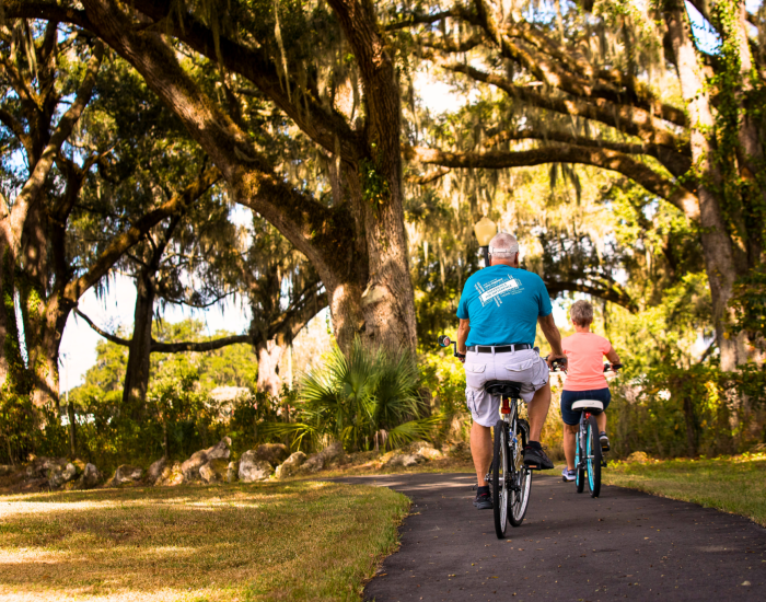 Two people ride bicycles on a paved path through a shaded park at an RV resort in Bushnell, Central Florida, enjoying a sunny day outdoors amid large trees draped in Spanish moss.