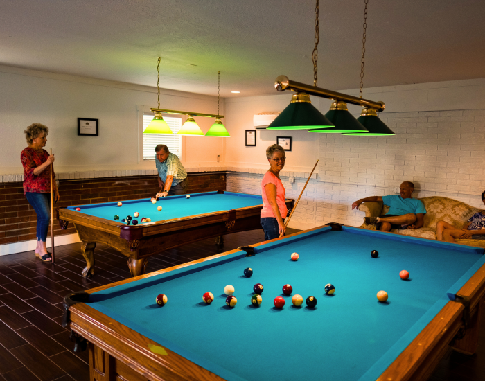 Five adults relax in the game room at Paradise Oaks RV in Bushnell. Two play pool while another stands close by, and two watch from a couch against the wall. Hanging lights illuminate the tables above the wood floors, creating a cozy RV resort vibe.