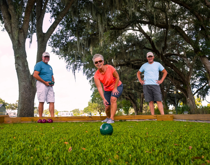 Three older adults play bocce ball on a grass court under shady trees at Paradise Oaks RV in Bushnell, Central Florida. One is mid-throw and smiling, while the others watch. The scene is bright and cheerful.