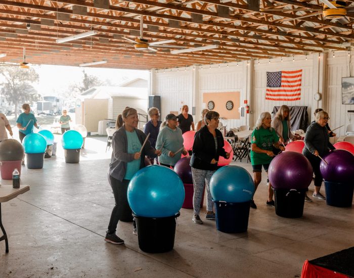 A group of people exercise indoors at an RV resort in Bushnell, Central Florida, using large colorful stability balls on buckets. They appear to be following a fitness class in a spacious building with an American flag on the wall.