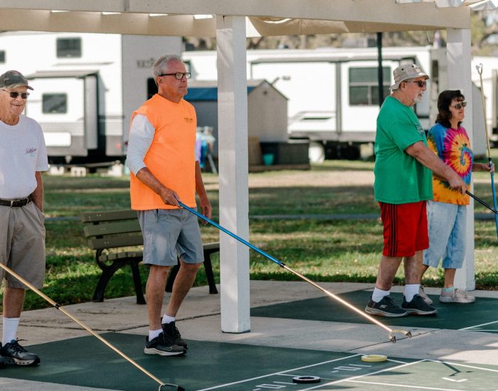Four older adults play shuffleboard outdoors at a Bushnell RV resort in Central Florida, each holding a cue stick. They stand on a shuffleboard court, smiling and engaged, with RVs and lush greenery in the background.