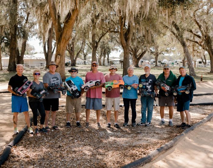 A group of eleven people stand outdoors on a dirt track at an RV resort in Bushnell, Central Florida, each holding a remote-controlled car. Trees with hanging moss are in the background, and everyone is smiling and dressed casually.