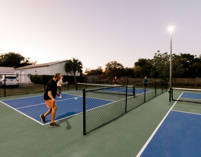 At dusk, four people play pickleball on outdoor courts at Paradise Oaks RV in Bushnell. Two players are in the foreground, facing their opponents across the net, with trees and buildings providing a scenic resort backdrop.