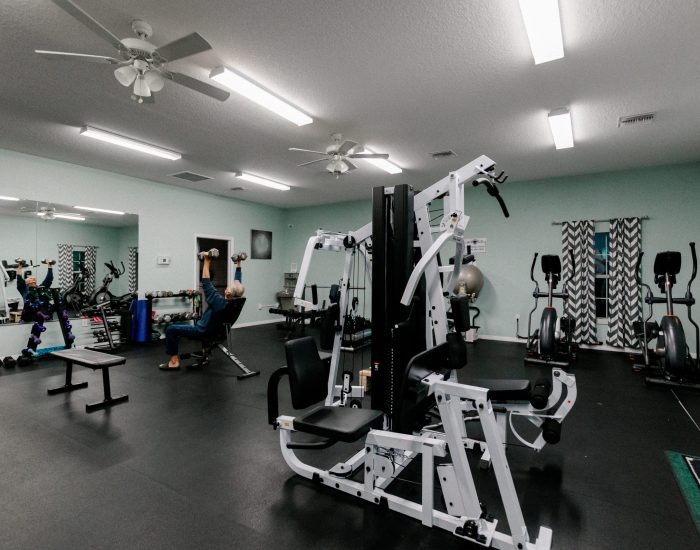 A spacious gym at Paradise Oaks RV Resort in Bushnell features various exercise equipment, including a weight machine, free weights, benches, and cardio machines. A person is seated and lifting dumbbells in front of a large wall mirror.