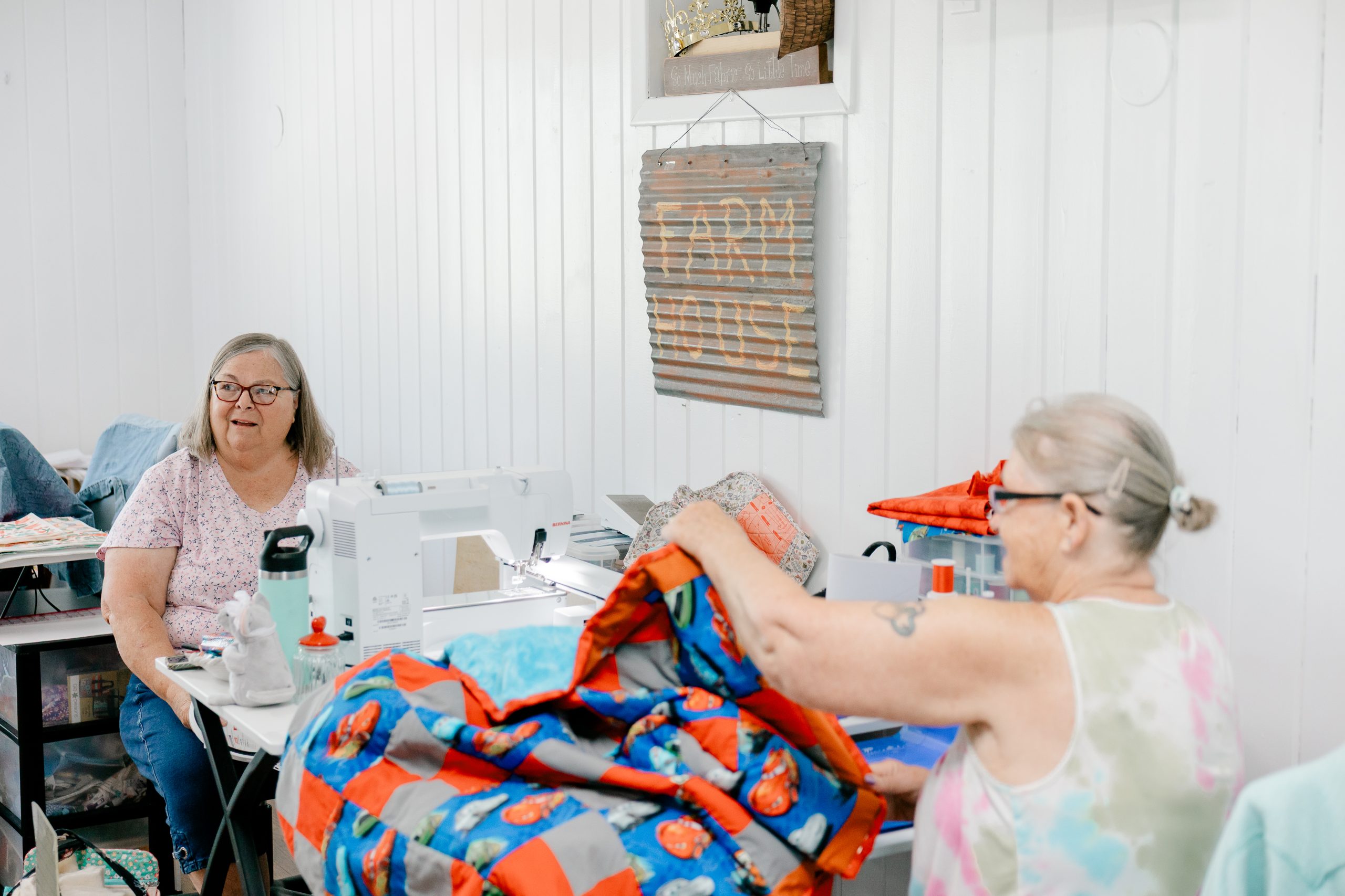 Two women sit at tables in a bright room working on colorful quilts, one using a sewing machine and the other folding fabric. A rustic Farm Fresh sign hangs on the white-paneled wall behind them. Two women sit at tables in a bright room working on colorful quilts, one using a sewing machine and the other folding fabric. A rustic Farm Fresh sign hangs on the white-paneled wall behind them.