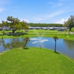 A small pond with a central water fountain is surrounded by green grass and palm trees, with RVs and buildings in the background under a clear blue sky.
