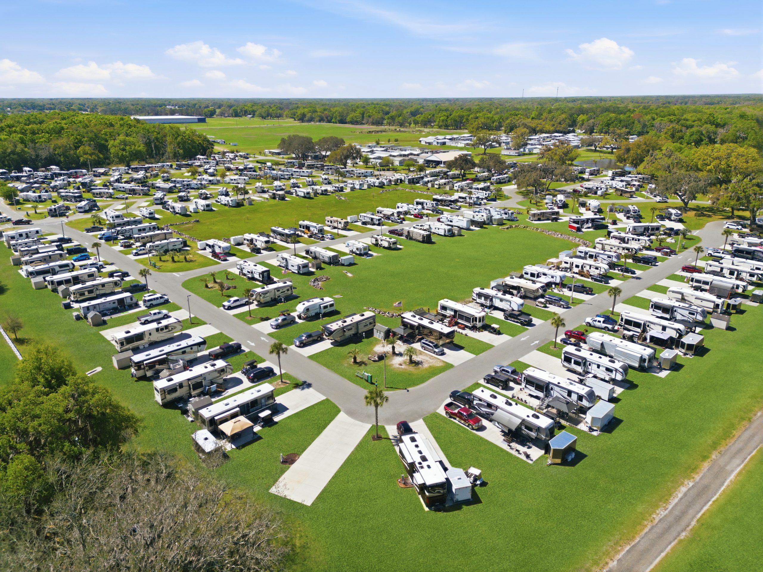 Aerial view of a large, organized RV park with numerous RVs parked in rows on grassy lots, intersected by paved roads, surrounded by green trees and open countryside under a blue sky with scattered clouds.