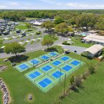 Aerial view of a recreational area with several blue and green pickleball courts, parked RVs, trees, open grassy fields, and nearby buildings under a clear blue sky.