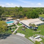 Aerial view of a recreational facility with a large building, outdoor swimming pool, golf carts, American flags, trees, grassy areas, and RVs parked nearby under a clear blue sky.