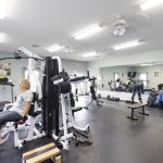 A woman uses a weight machine in a well-lit gym with various exercise equipment, mirrors on one wall, and free weights neatly arranged on racks.