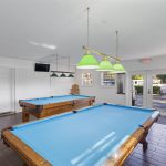 A bright game room with two blue pool tables, wooden floors, green hanging lights, white brick walls, a TV mounted high, and windows letting in natural light.