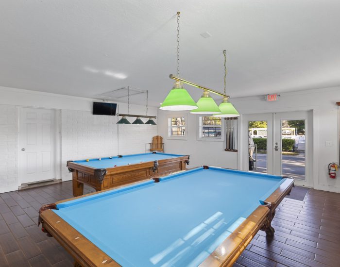 A bright game room with two blue pool tables, wooden floors, green hanging lights, white brick walls, a TV mounted high, and windows letting in natural light.