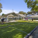 A group of people play bocce ball on an outdoor court under sunny skies, with a covered pavilion and trees in the background. Some people are standing and watching while others are actively playing.