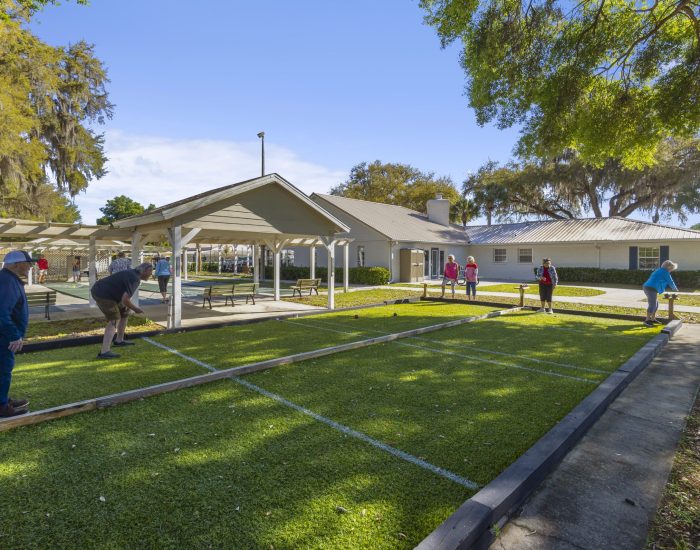 A group of people play bocce ball on an outdoor court under sunny skies, with a covered pavilion and trees in the background. Some people are standing and watching while others are actively playing.