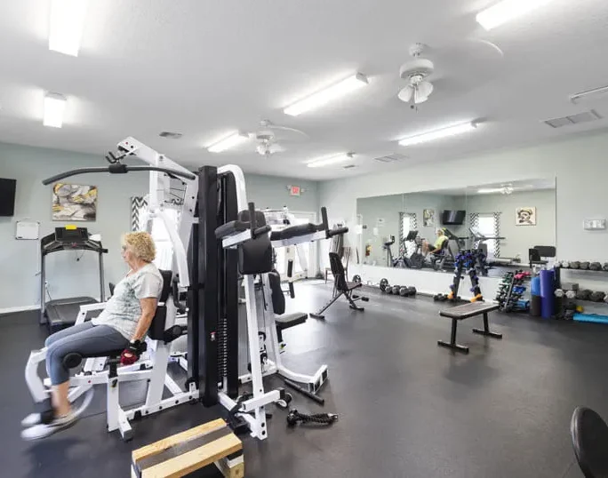 A woman exercises on a weight machine in a well-lit gym with various equipment, including free weights, benches, mirrors, and a treadmill. The room has white walls and ceiling lights.