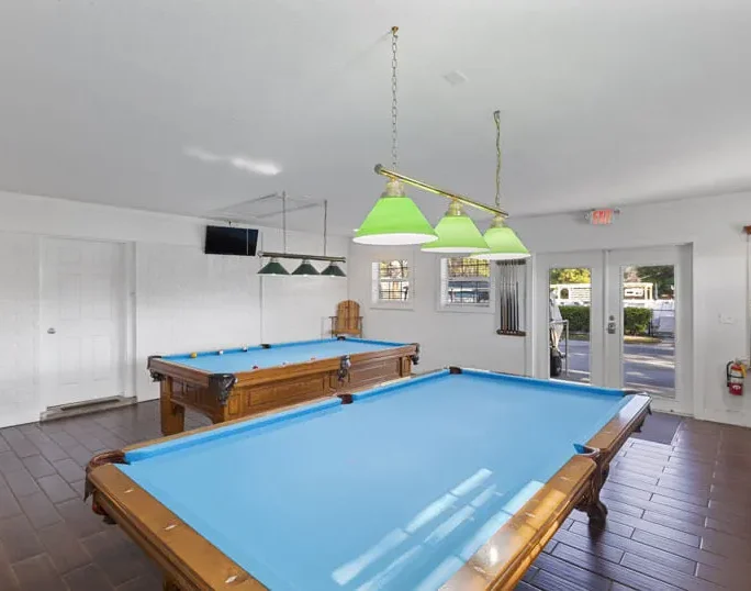 Two pool tables with bright blue felt sit in a spacious, well-lit game room with wood-look flooring, green overhead lights, a TV, and pool cues on a wall rack. Large windows let in natural light.