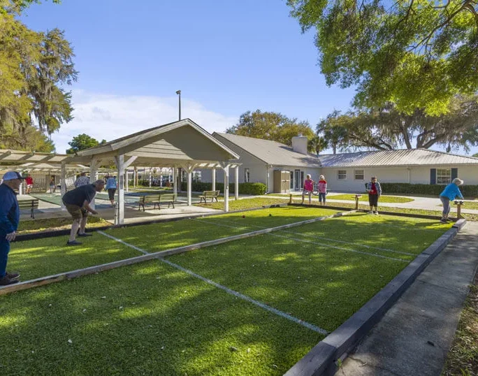 People play bocce ball on a grassy court outside near a covered pavilion, with trees and a building in the background under a clear blue sky.