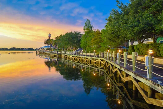 A wooden boardwalk lined with trees runs alongside a calm lake at sunset, with golden light reflecting on the water and a small lighthouse visible in the background.