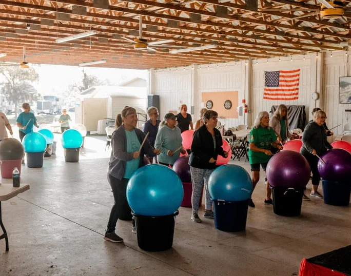 A group of adults exercise indoors, standing in front of large colorful stability balls placed on bins. The setting is a spacious hall with an American flag on the wall and sunlight streaming in.