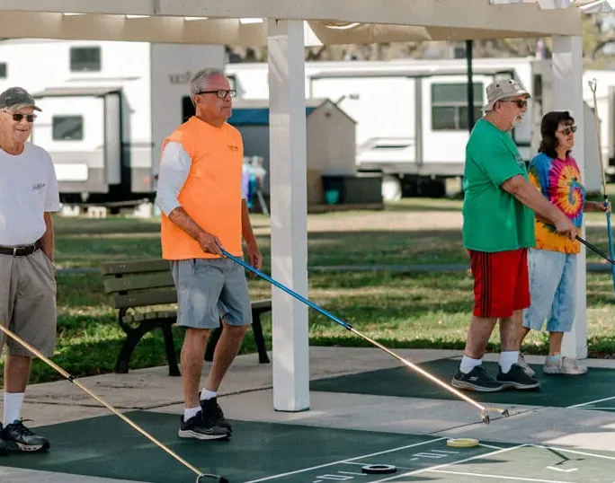 Four adults play shuffleboard outdoors. Three men and one woman stand in a row, holding cues. They are casually dressed and appear to be enjoying a sunny day at a recreational park with RVs in the background.