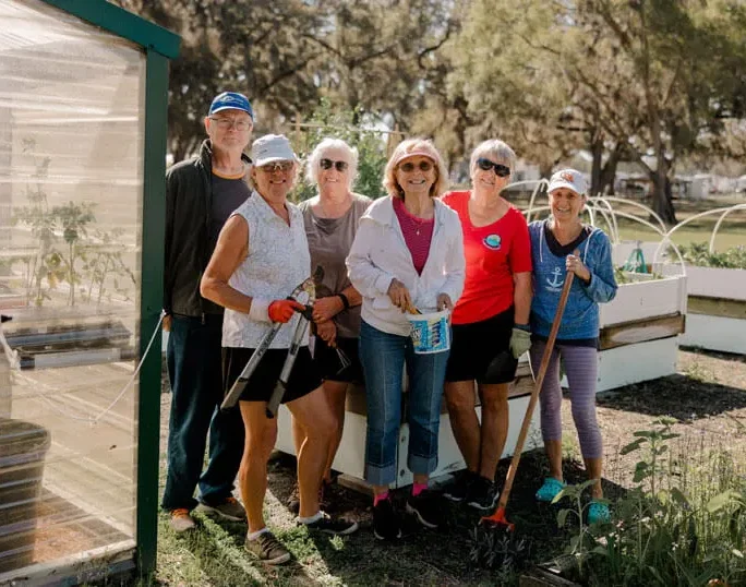 Six older adults stand together smiling in a community garden, holding gardening tools. Raised beds with plants and a greenhouse are visible, with trees in the background on a sunny day.