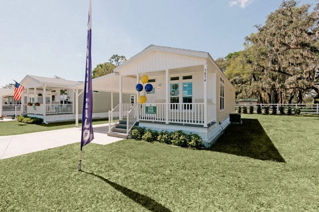 A small, white, modern manufactured home with a front porch decorated with yellow and blue balloons. The lawn is neatly trimmed, and an American flag is visible on a neighboring house. A tall sign stands in the grass.