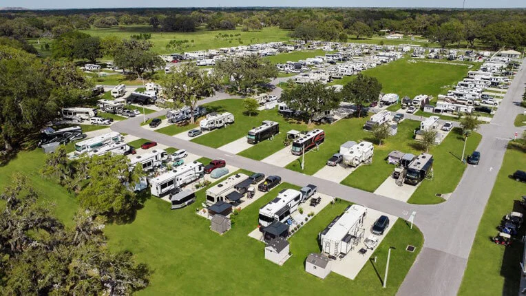 Aerial view of a large RV park with numerous recreational vehicles parked on individual lots, surrounded by grassy areas and trees, with roads dividing the sections.